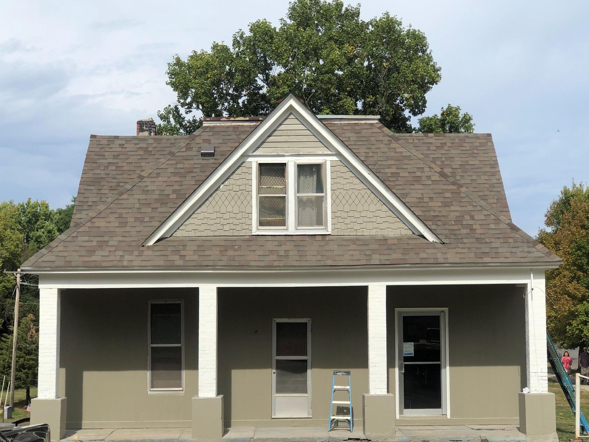 Tan house with a porch and gabled roof, tan siding, white trim, and a tree in the background.