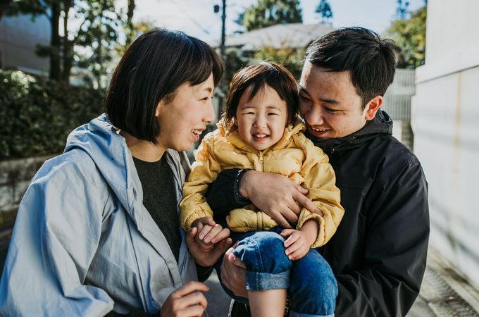 Family smiling, outdoors. Mother, father holding a child wearing yellow jacket.