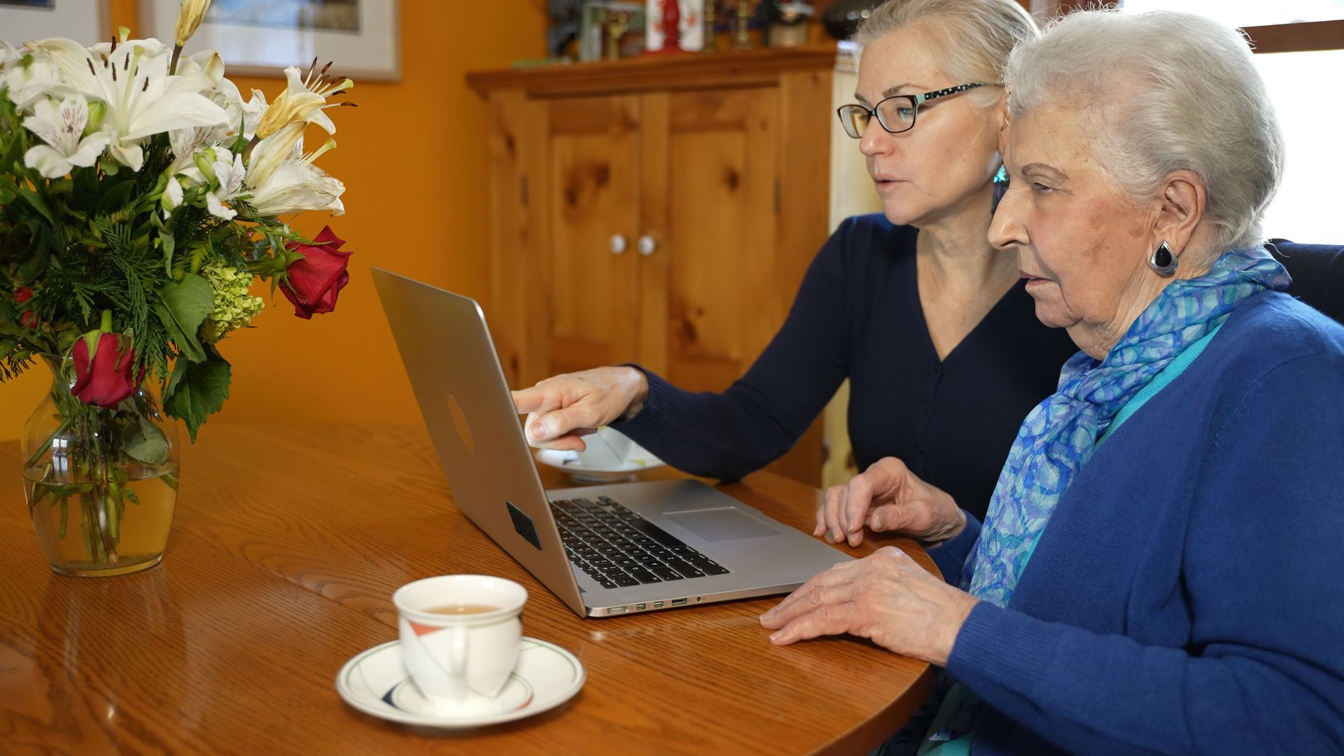 Woman helps elderly woman with laptop at a table, pointing at the screen. Coffee and flowers nearby.