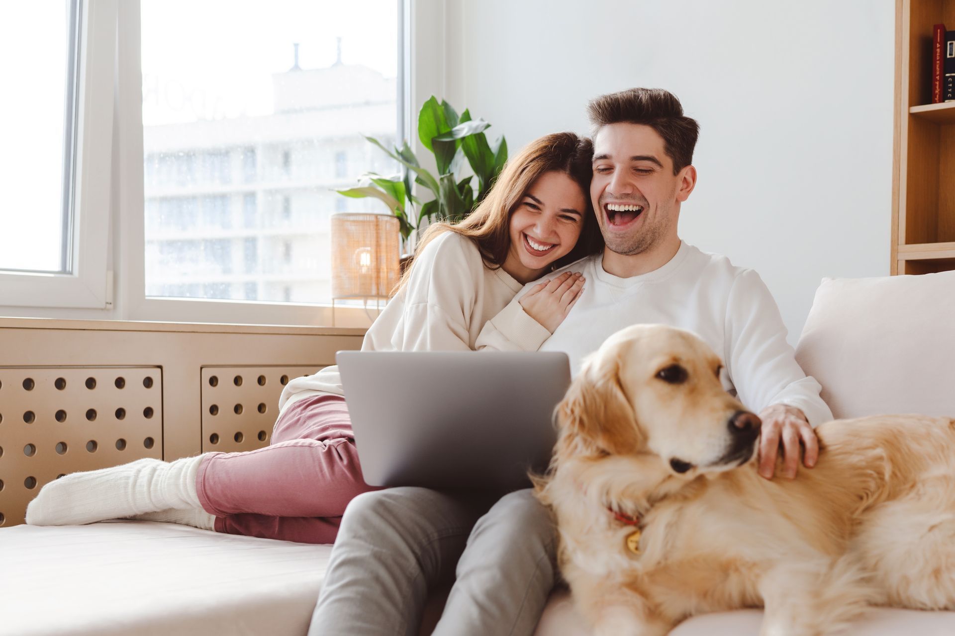Couple laughing on couch with a golden retriever and laptop in a bright living room.