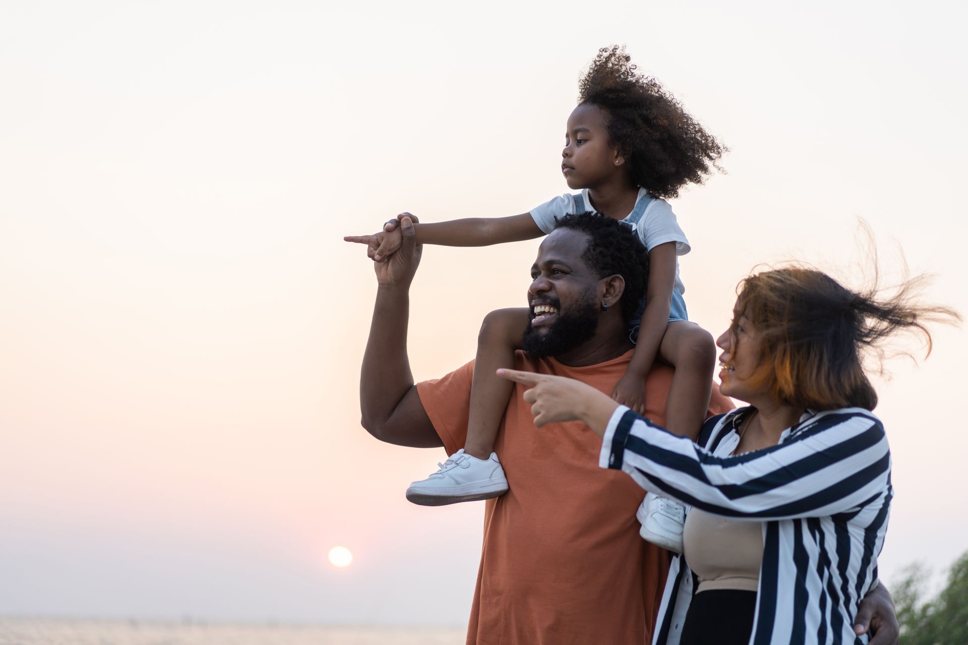 Family at sunset; father carrying child on shoulders, mother points.
