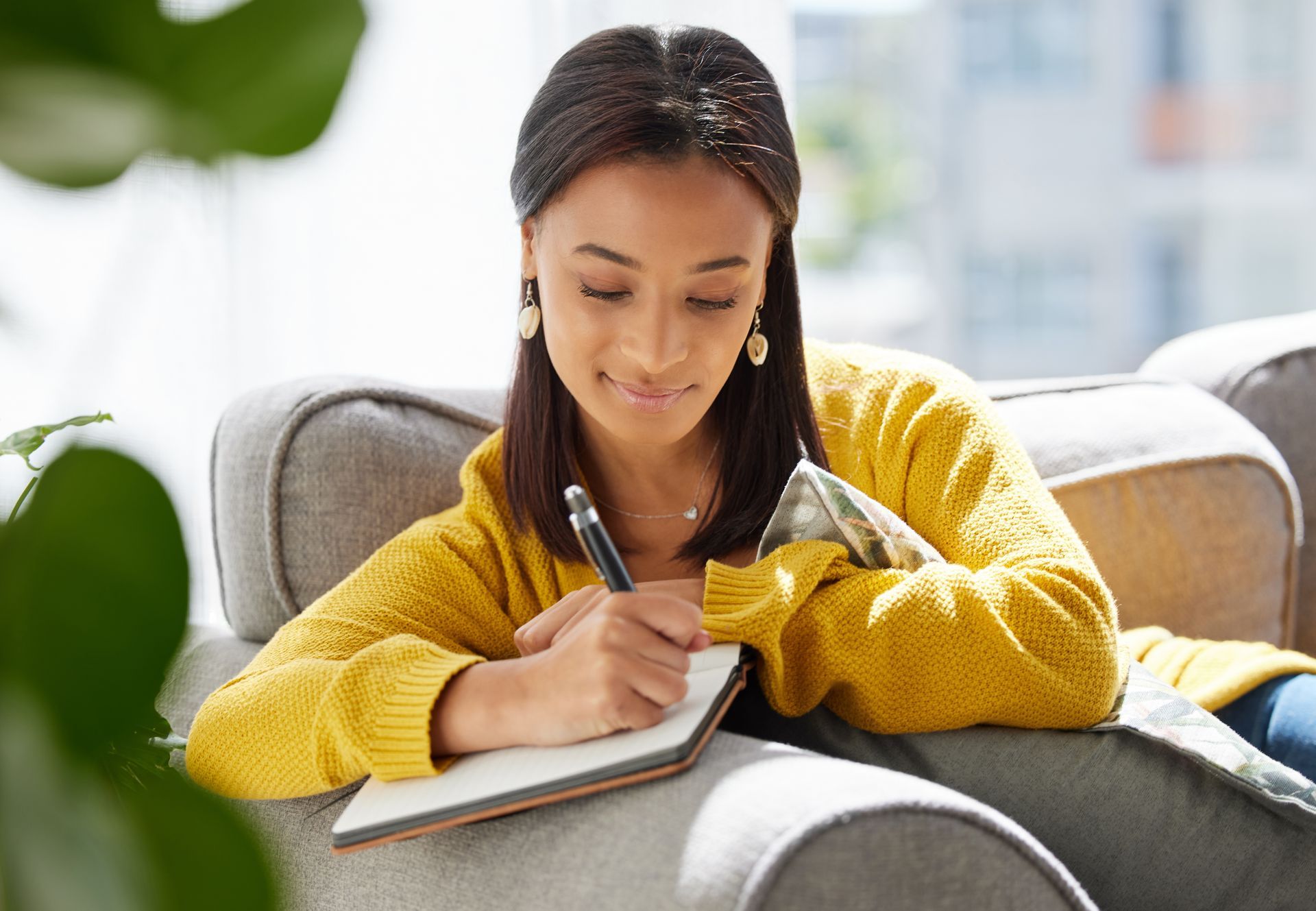 Woman writing in a notebook on a couch, wearing a yellow sweater.