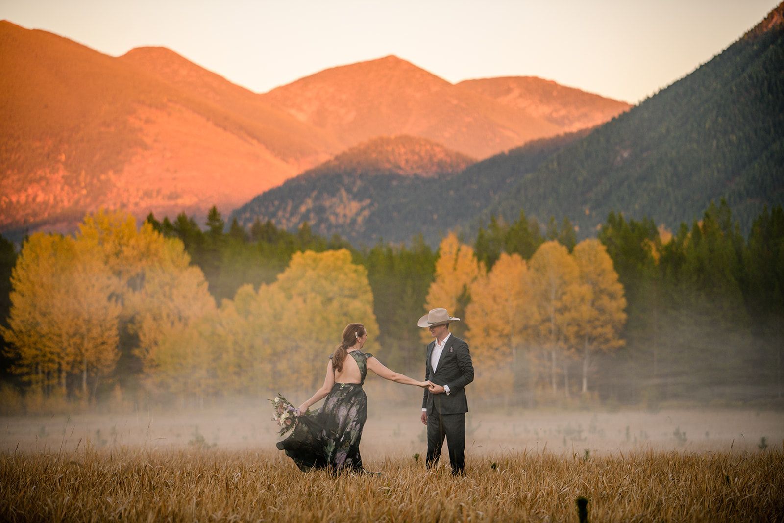 A bride and groom are dancing in a field with mountains in the background.