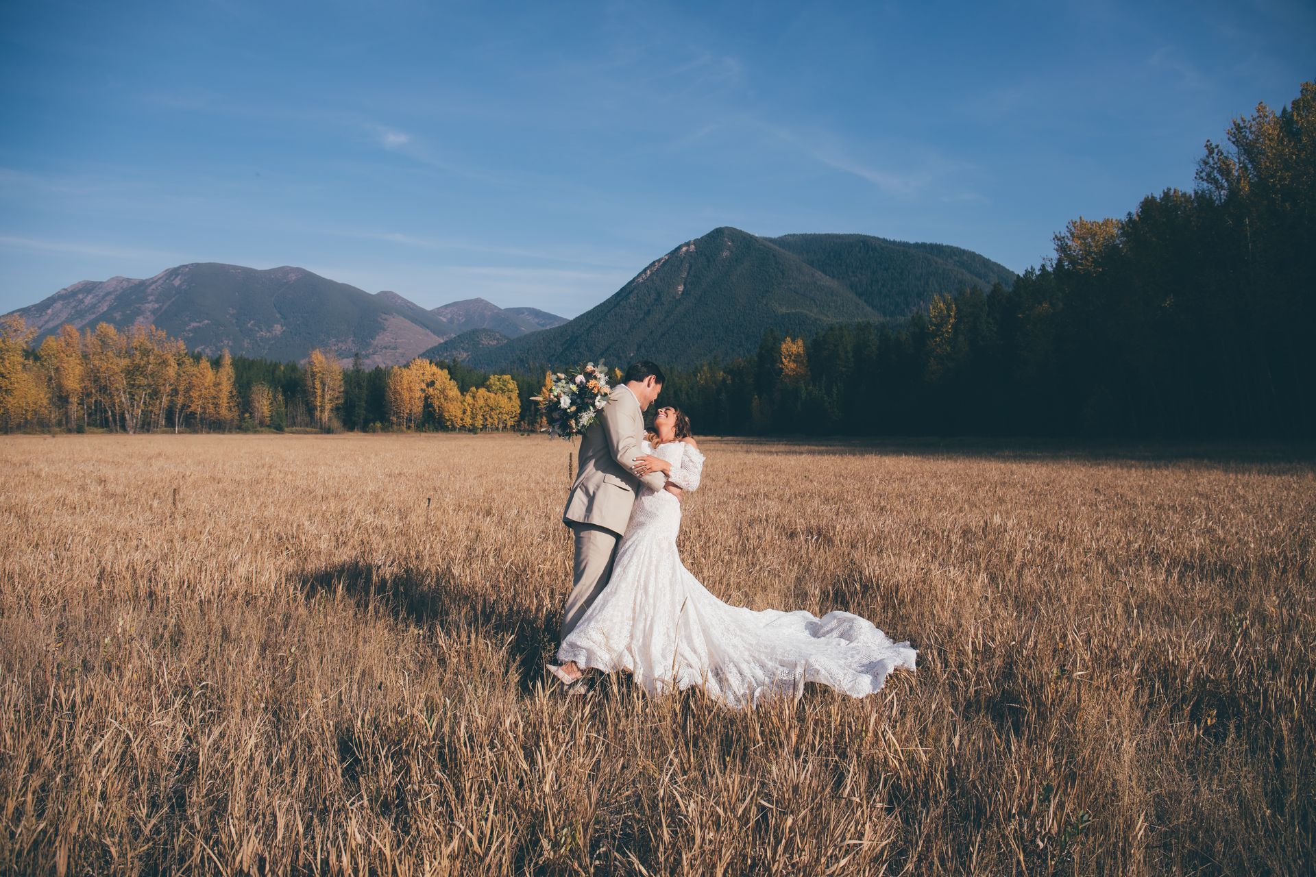 A bride and groom are standing in a field with mountains in the background.