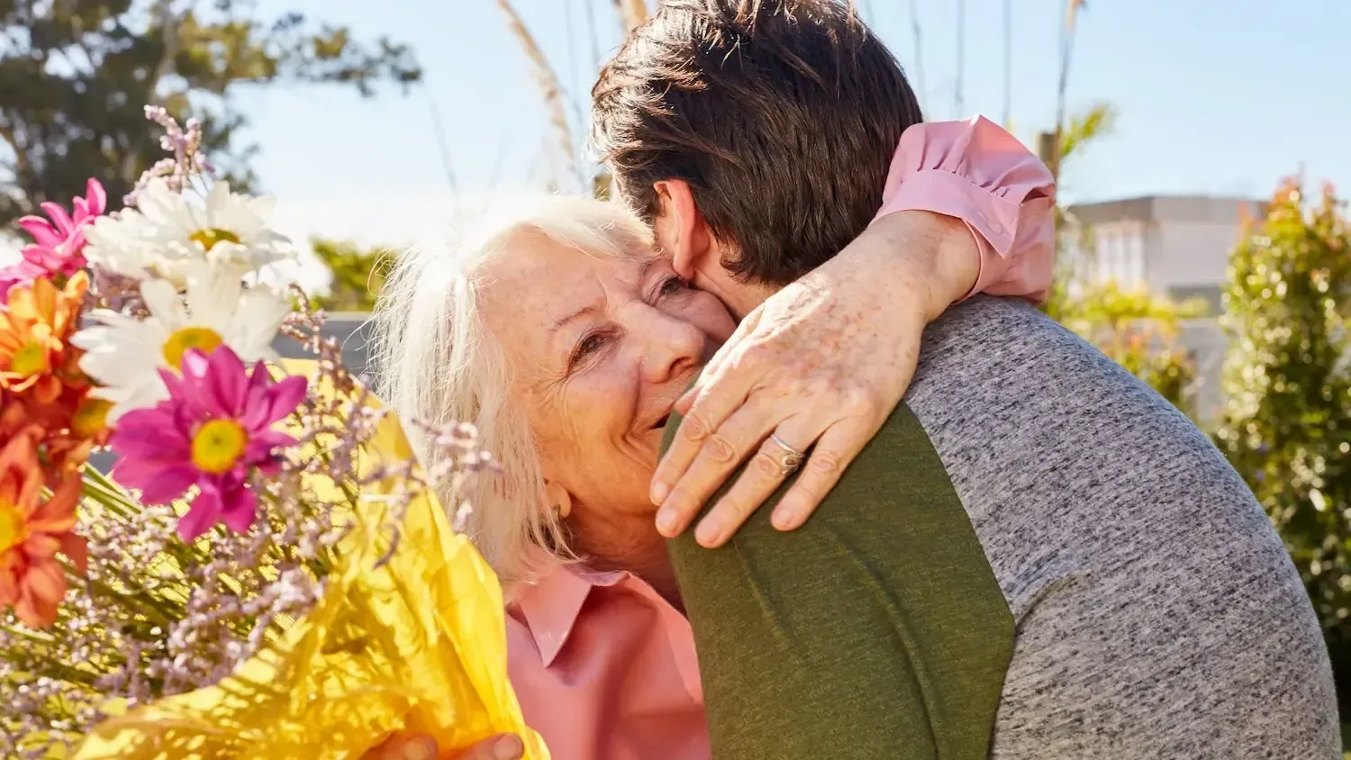 A man is hugging an older woman who is holding a bouquet of flowers.