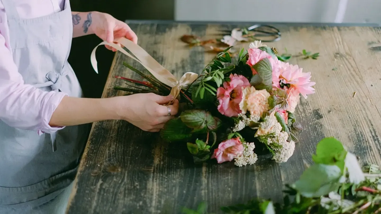 Person tying a ribbon around a bouquet of pink and white flowers on a wooden table, preparing for a floral arrangement.