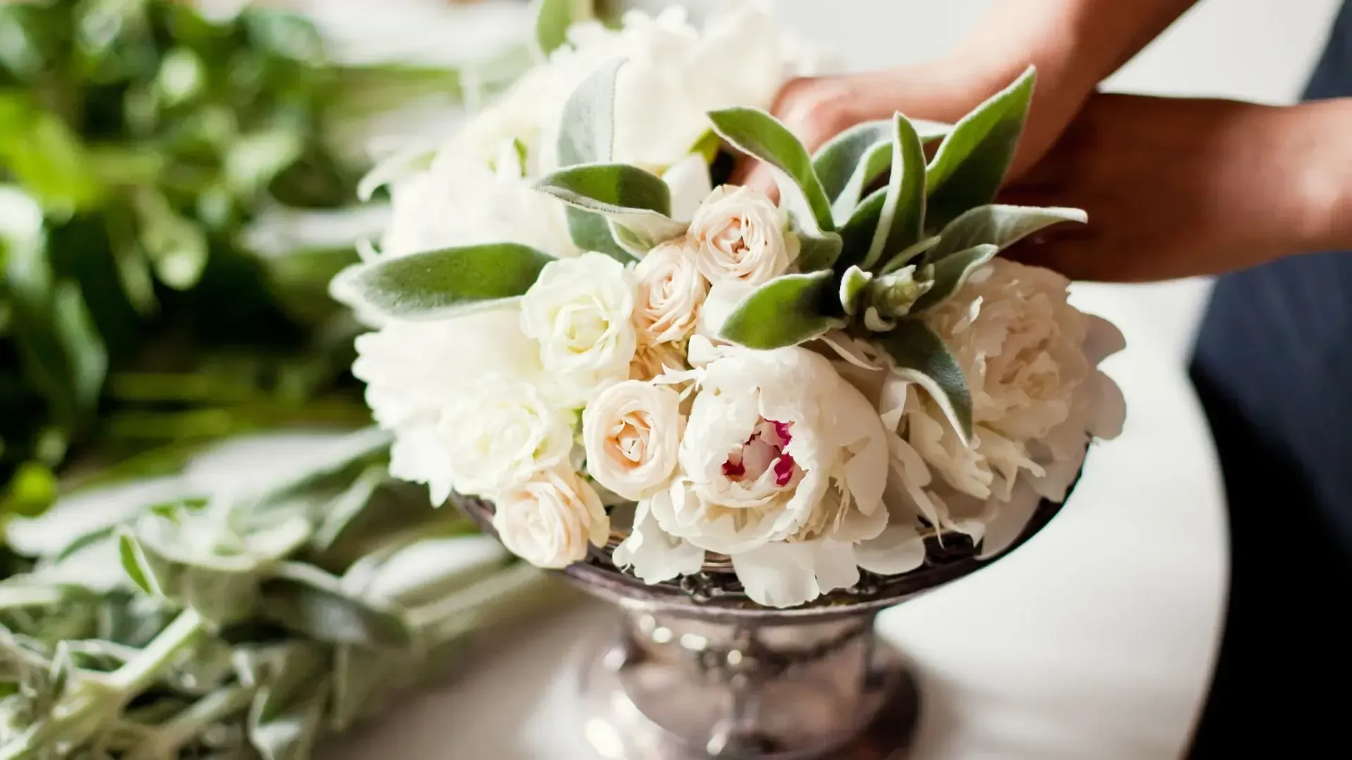 Person arranging a bouquet of white and peach flowers, with greenery, in a silver vase.