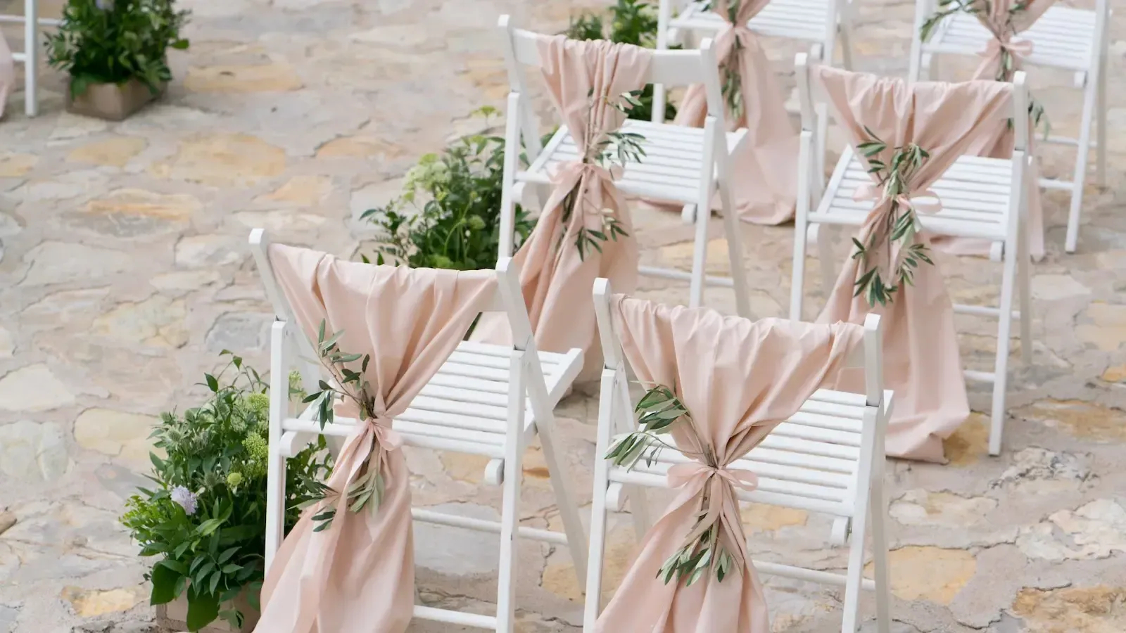 White chairs with draped blush fabric and greenery, arranged for an outdoor event on stone.