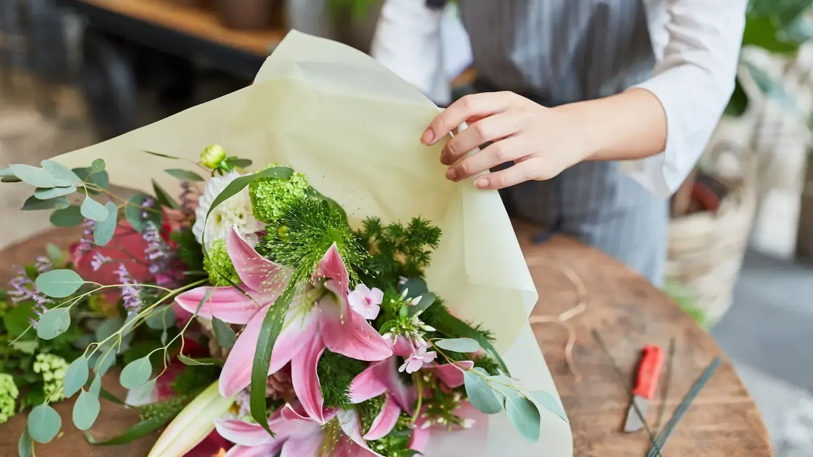 Person wrapping a bouquet of pink lilies and greenery with light green paper on a wooden table.