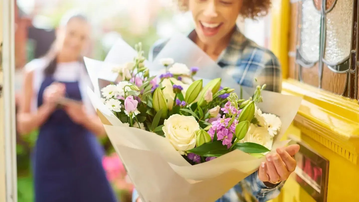 Woman receiving a bouquet of flowers at her door; smiling, shop worker in background.