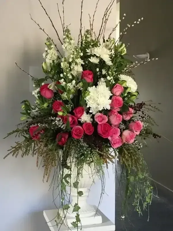Floral arrangement in a white urn. Pink roses, white flowers, greenery, and branches against a gray wall.