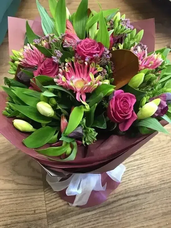 A bouquet of pink flowers wrapped in burgundy paper is sitting on a wooden table.