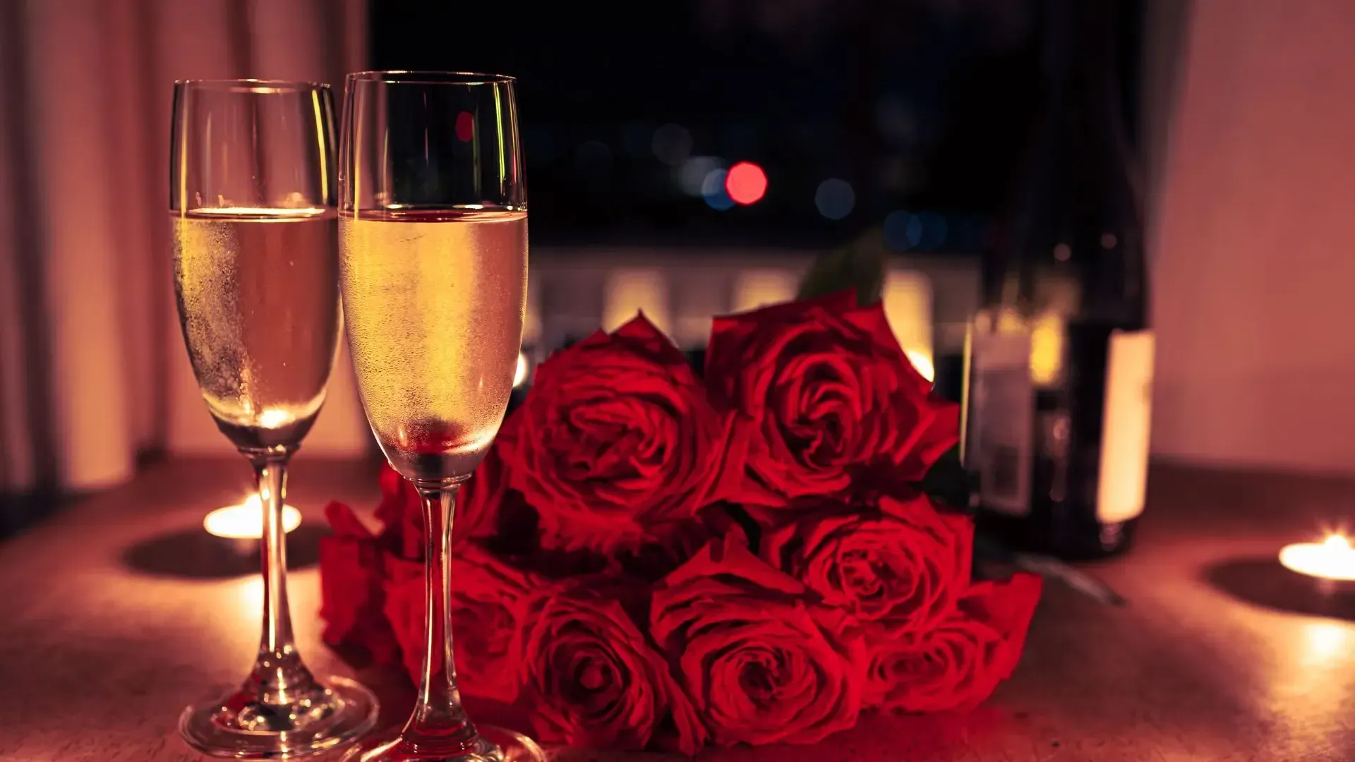 Two champagne flutes and red roses, romantic table setting with candles and bottle.