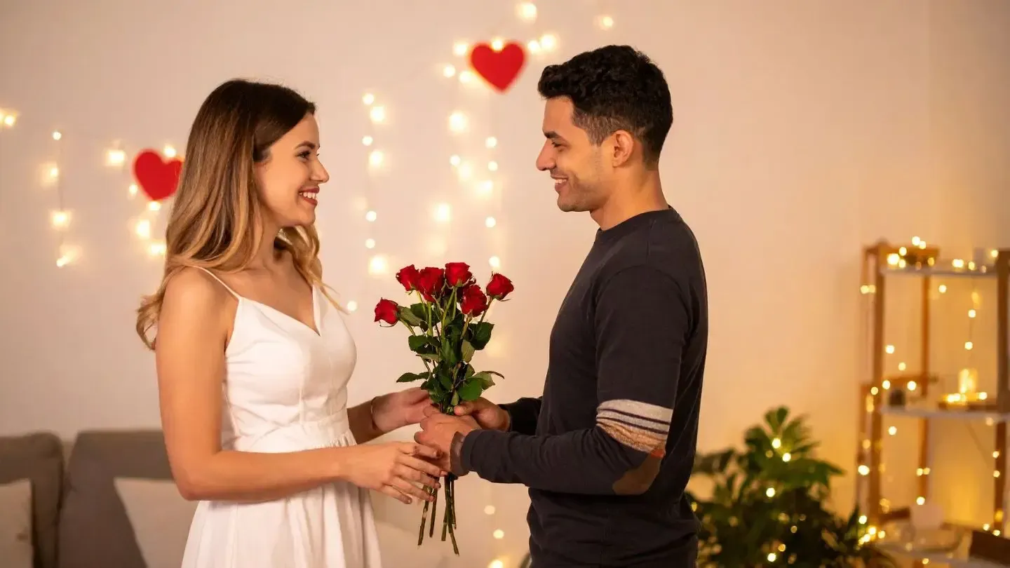 Man giving a woman red roses. They smile at each other indoors, with string lights and heart decorations.