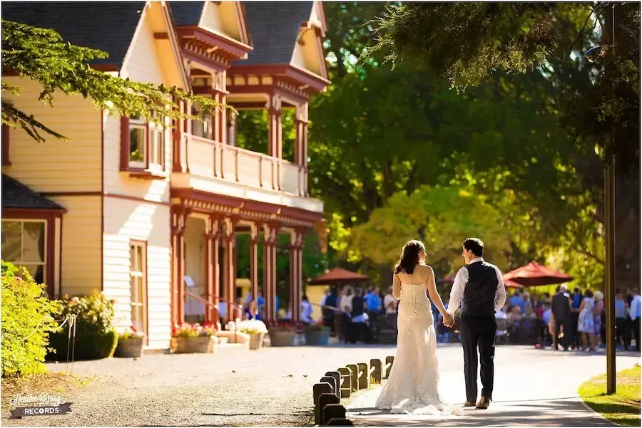 Bride and groom walk away from a Victorian house toward a crowd of people.