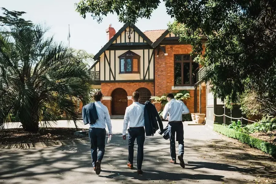 Three men in formal attire walk toward a brick Tudor-style building.