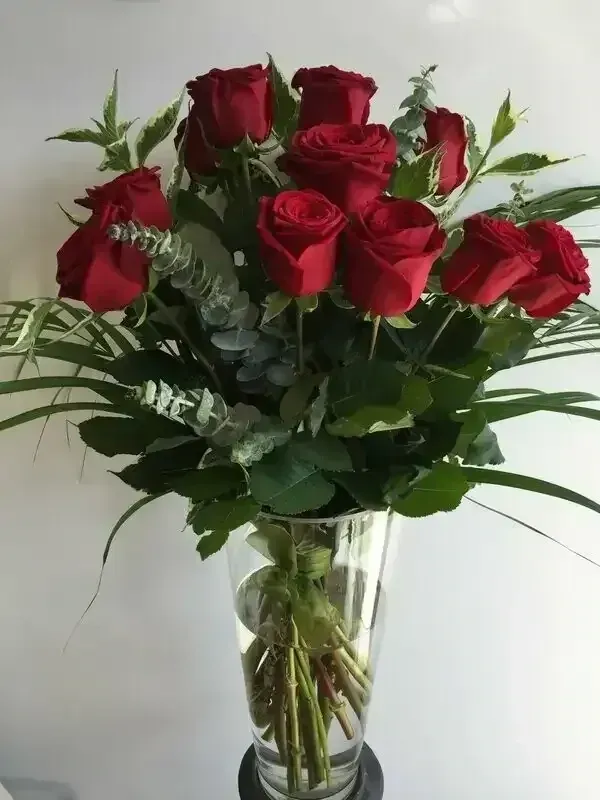 Red roses in a clear glass vase, surrounded by greenery against a white background.