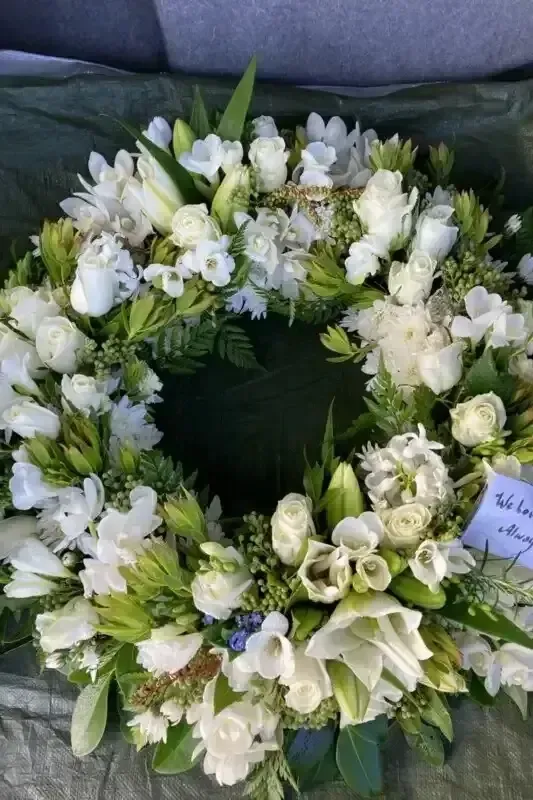 White floral wreath with lilies, roses, and other greenery on a green surface.