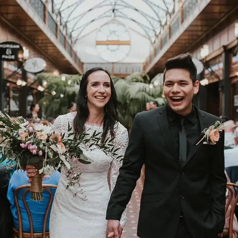 Newlyweds walking happily in a decorated arcade, bride in white dress holding flowers, groom in black suit.