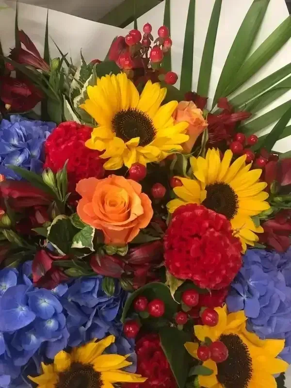 A vibrant floral bouquet featuring yellow sunflowers, orange roses, red carnations, blue hydrangeas, and red berries.