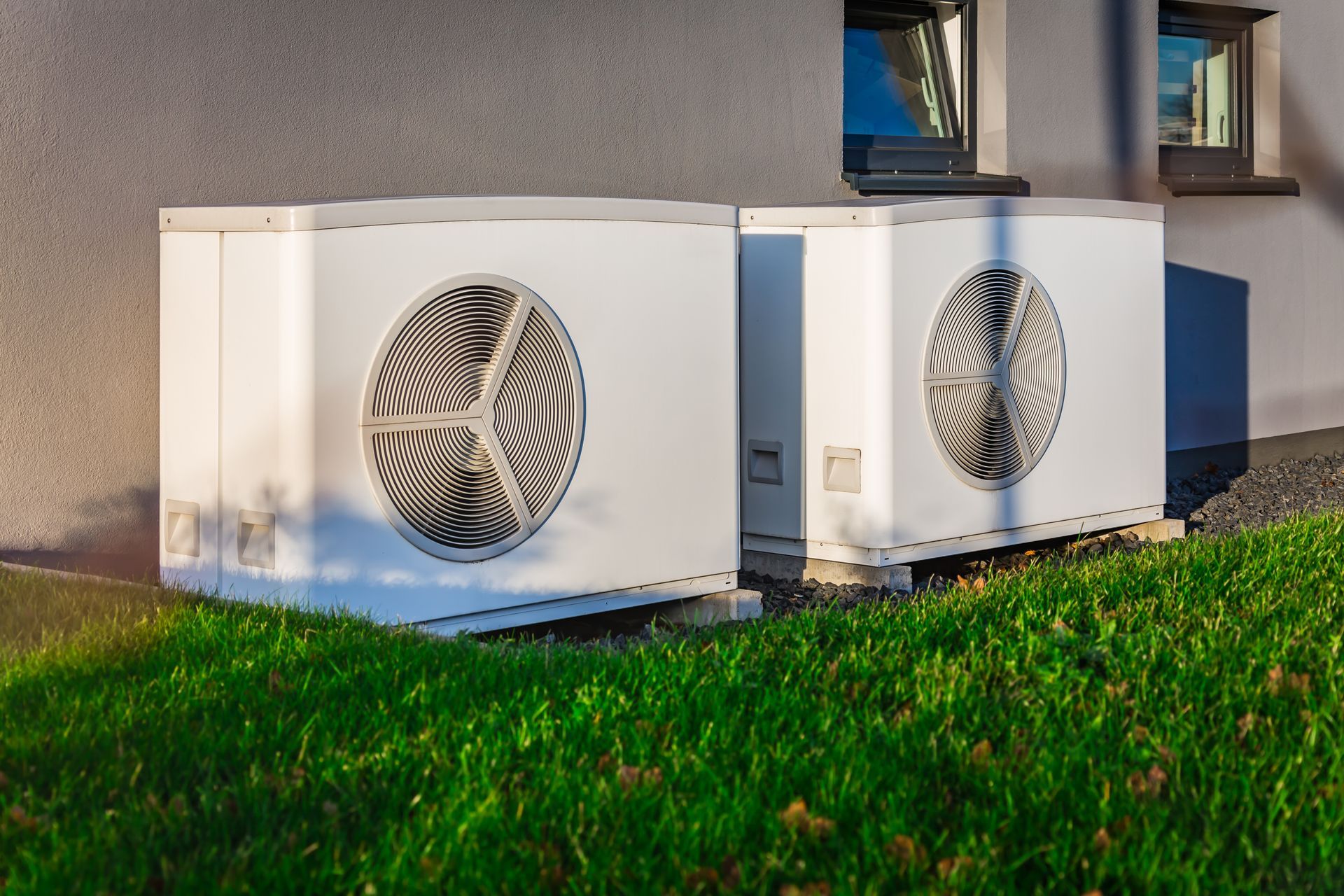 Two white heat pump units on a patch of green grass beside a building with dark-framed windows.