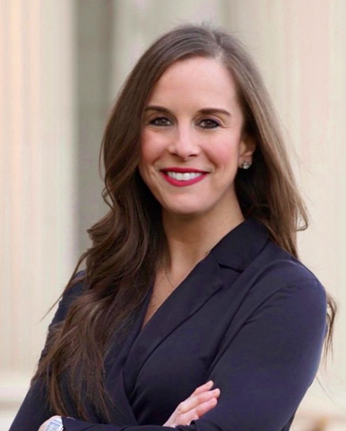 A person with long brown hair smiling with arms crossed, wearing a dark navy blazer, standing in front of a white column.