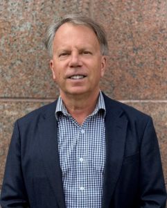 A person with light hair wearing a dark blazer over a blue and white checkered shirt, standing before a stone wall.