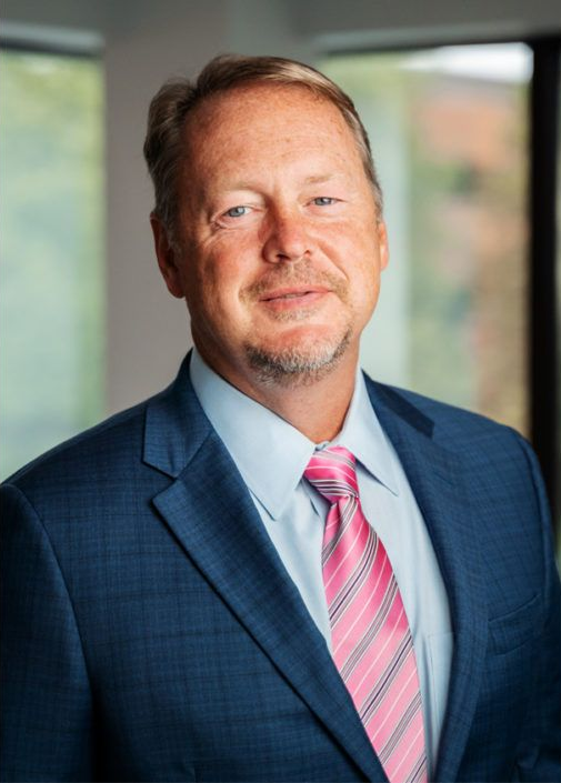 A professional headshot of a person wearing a navy blue suit, light blue shirt, and a pink and white striped tie.