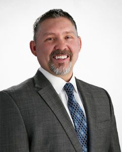 A person smiling, wearing a gray suit, white collared shirt, and a blue patterned tie against a plain white background.