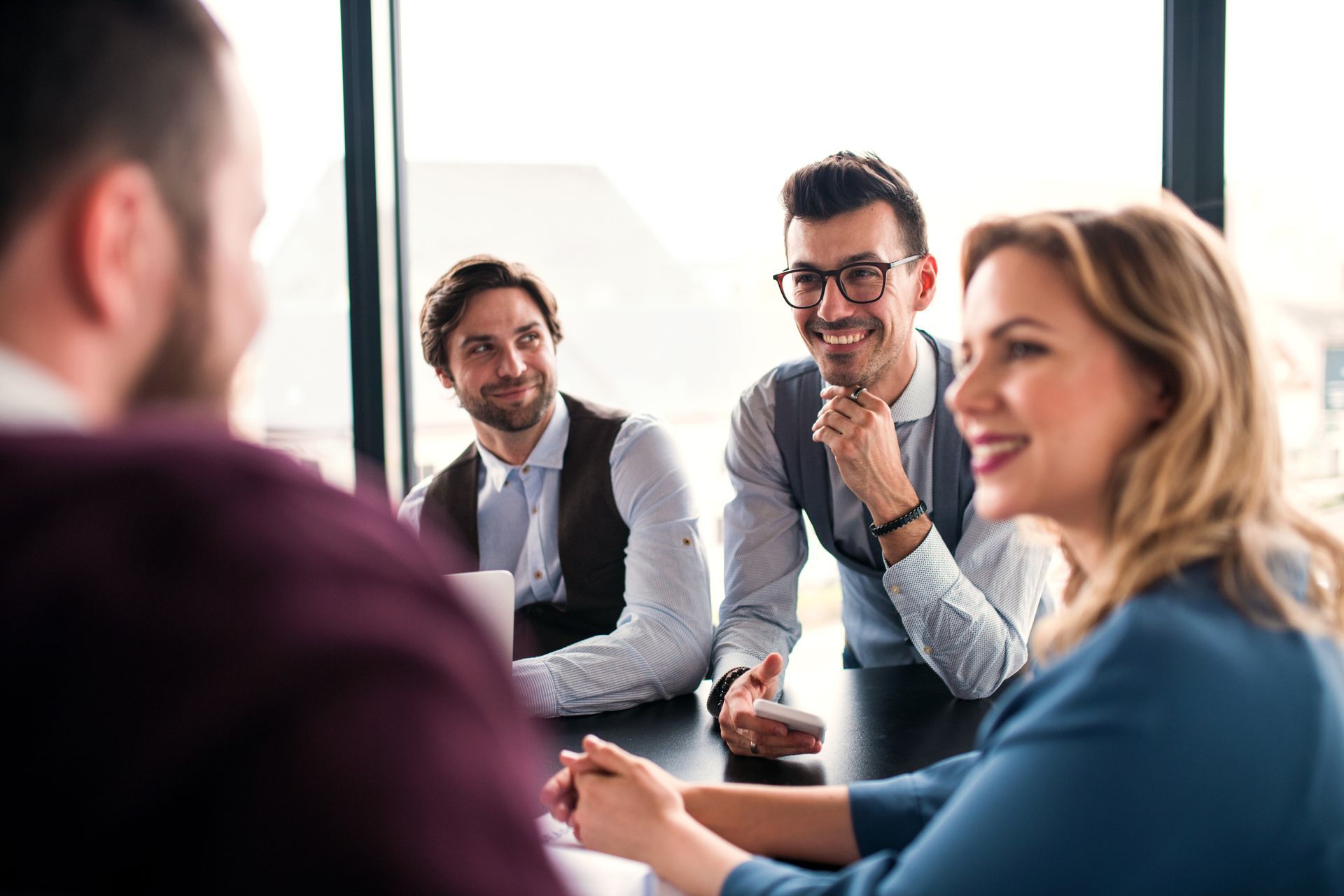 Four people in business attire sit around a table in a sunlit office, engaged in a discussion with smiles.