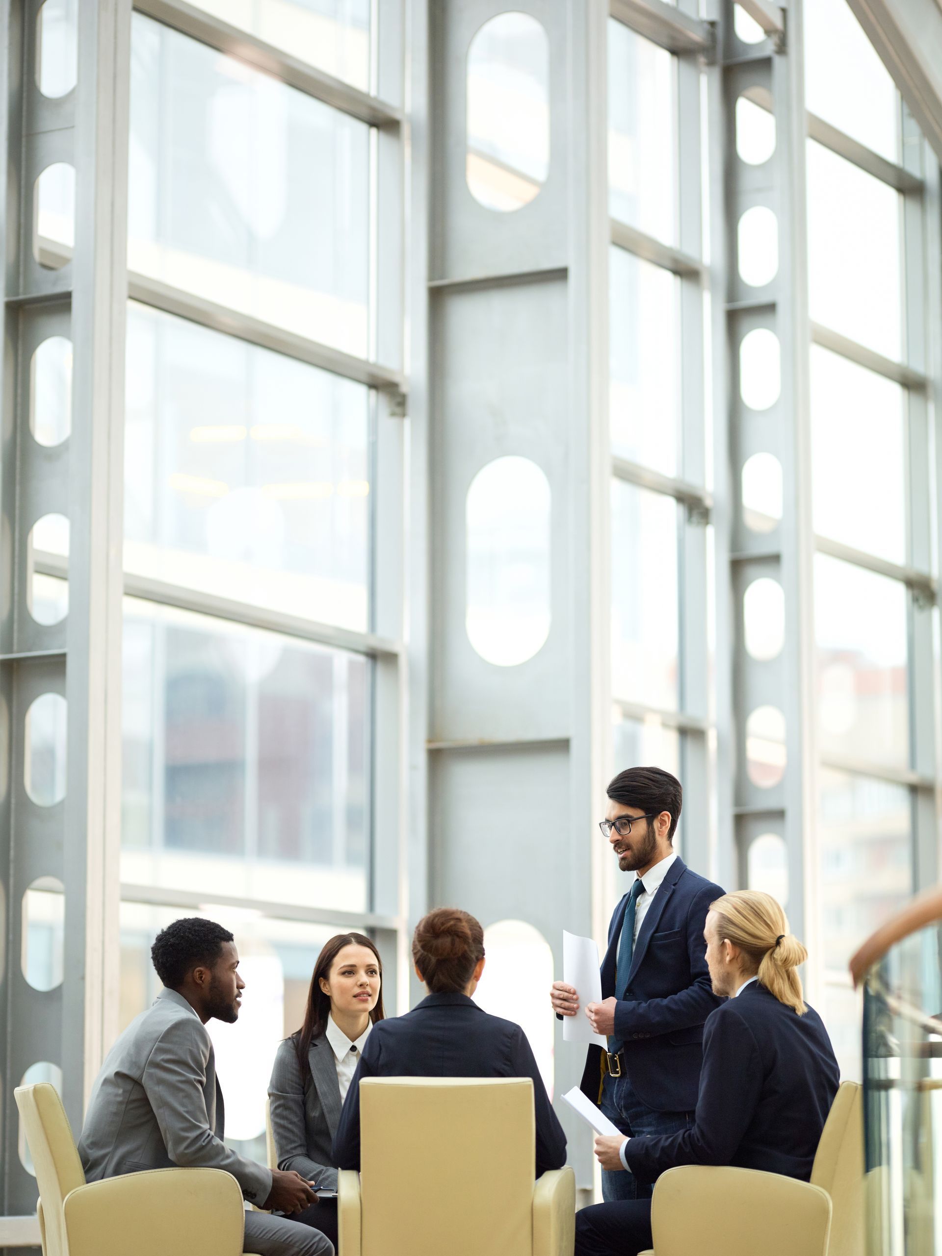 A group of four business professionals in suits discuss work while sitting in chairs in a modern office with large windows.