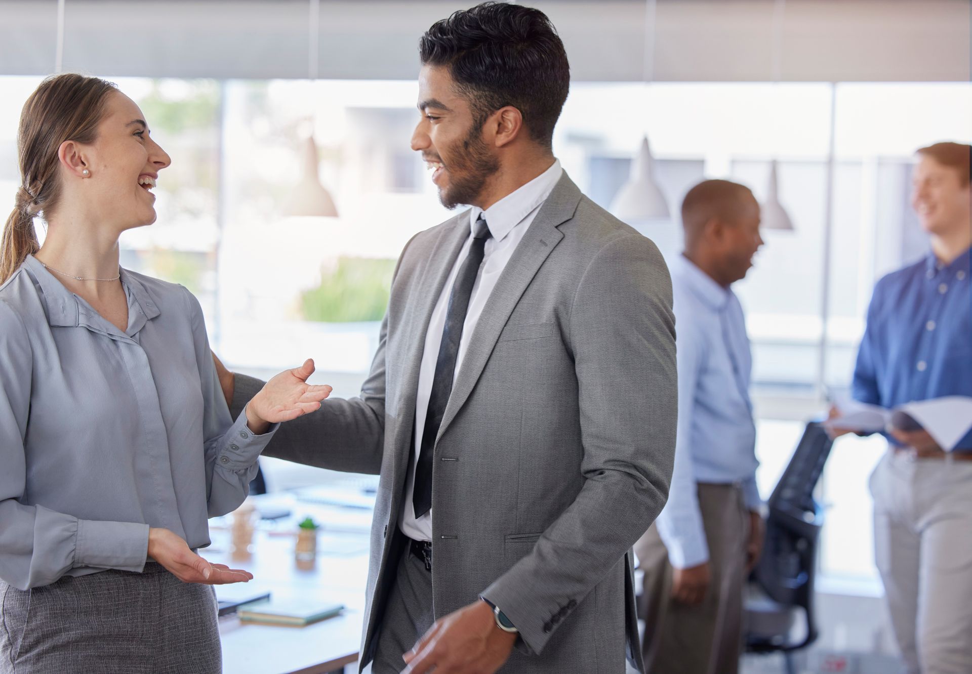 Colleagues smile and converse in a bright, modern office as others interact in the background.