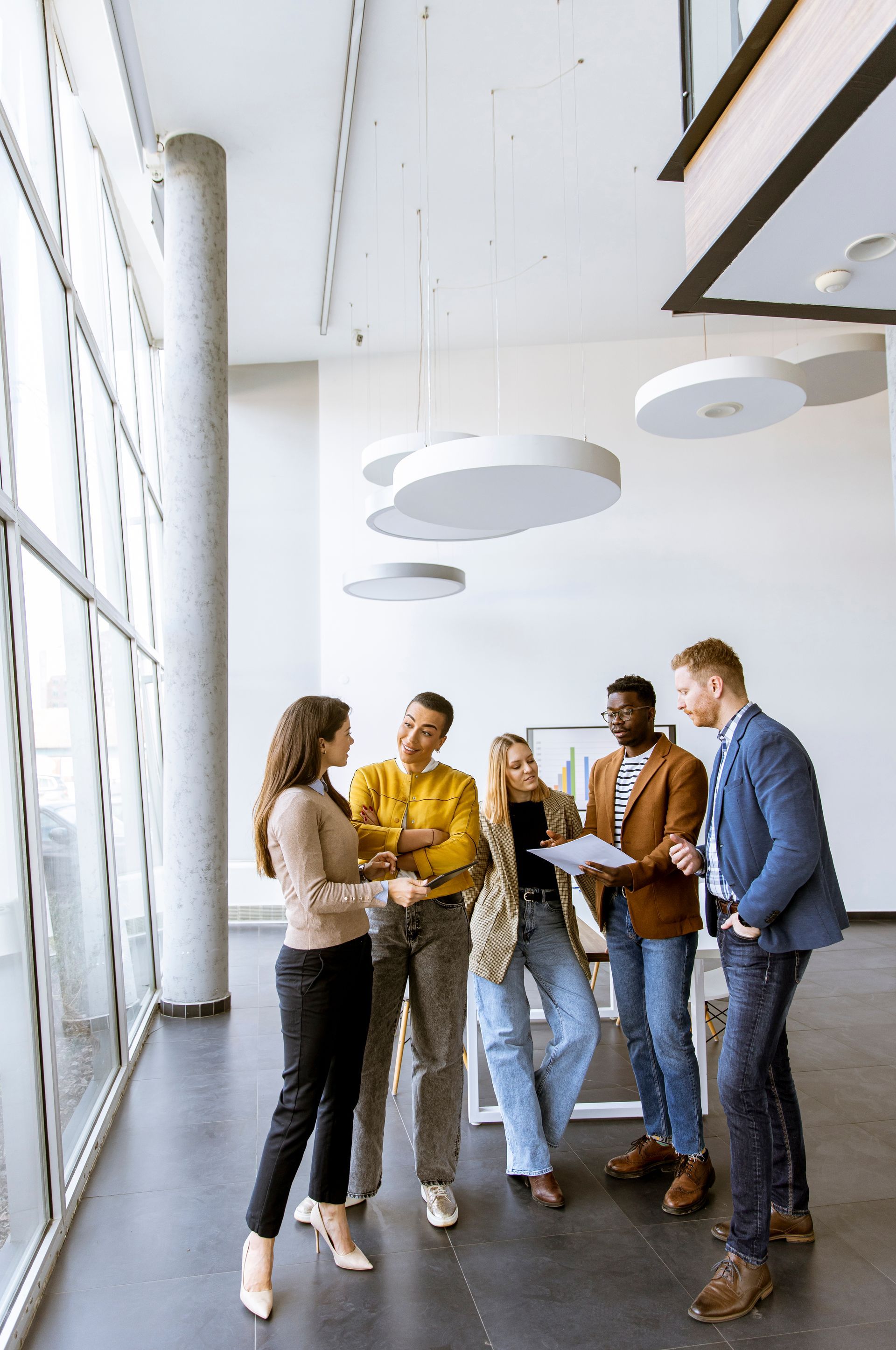 A group of five people in business casual attire stand in a bright, modern office, discussing documents together.
