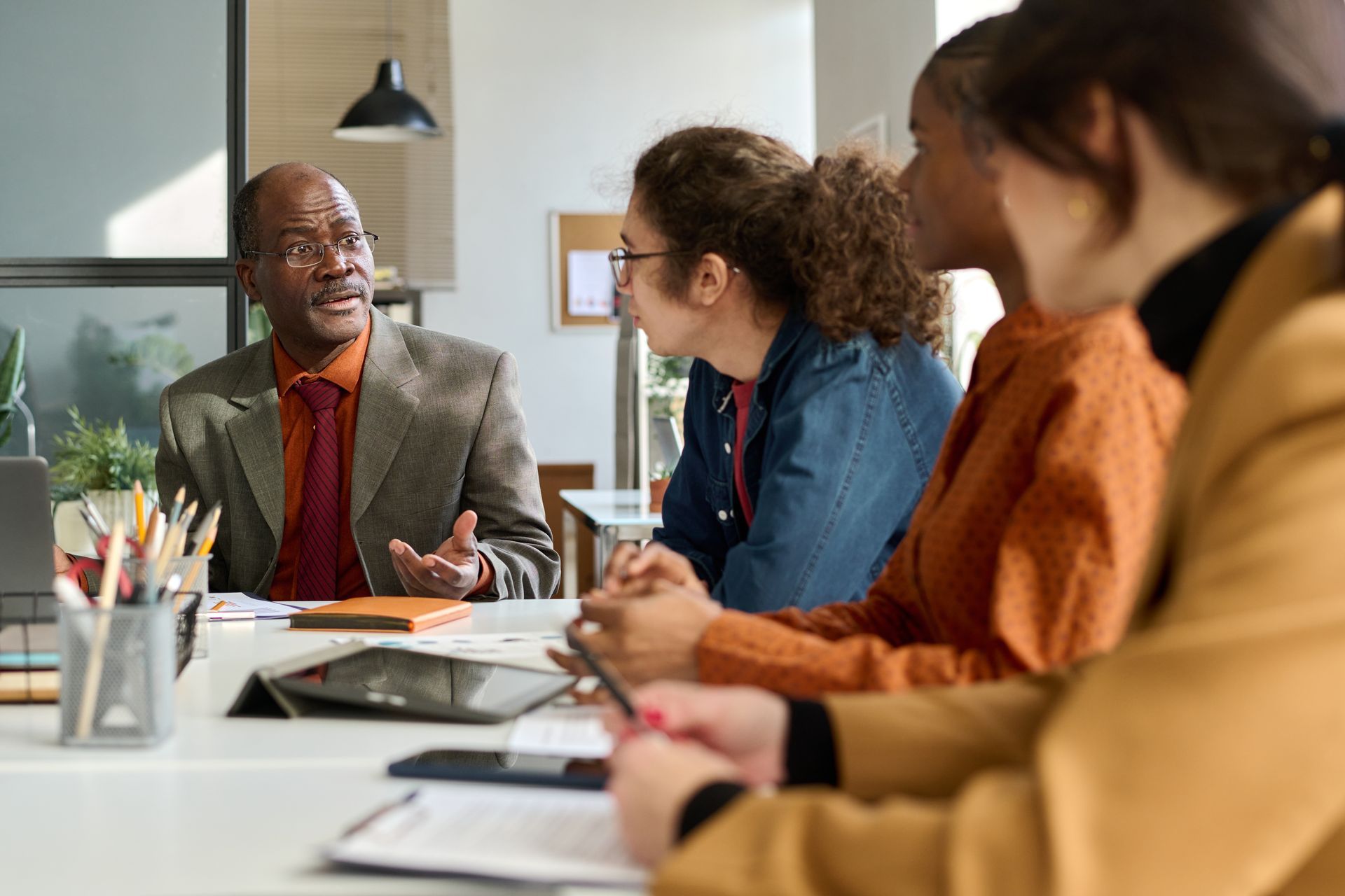 Four people sit around a meeting table in an office, with one person speaking while gesturing to the others.