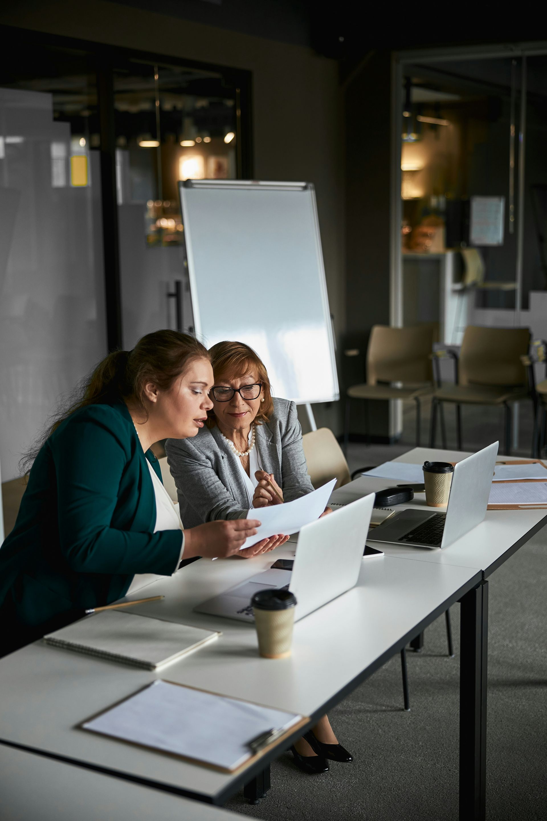Two colleagues sit at a white conference table in an office, reviewing documents and working on laptops.