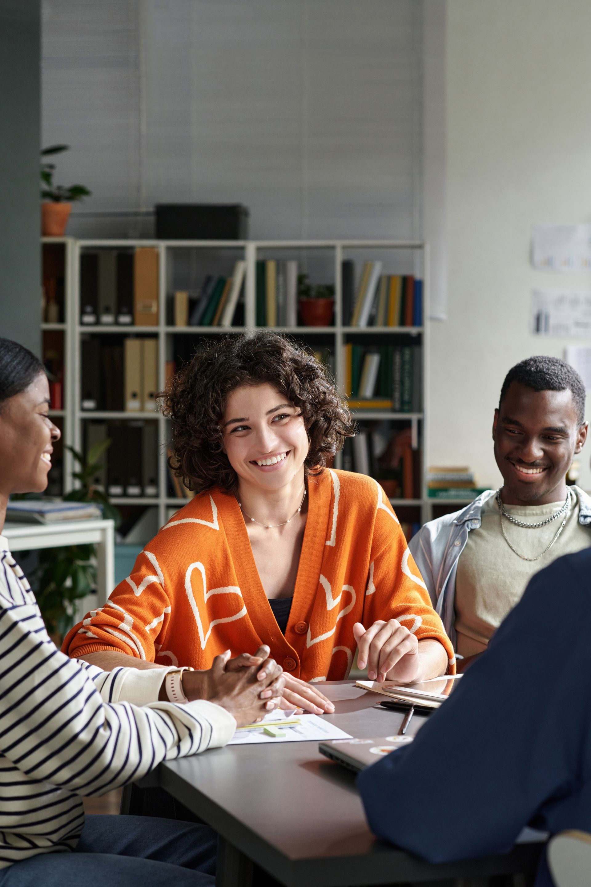 Four people sit around a table in an office, laughing and engaging in a collaborative discussion near a bookshelf.