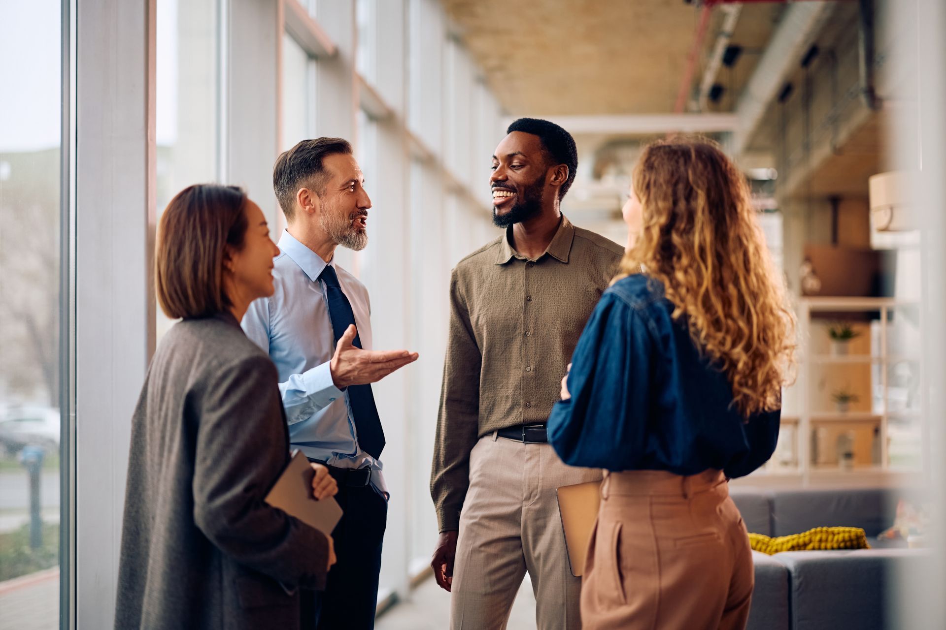 Four professionals in business casual attire stand in a sunlit office hallway, smiling and chatting.