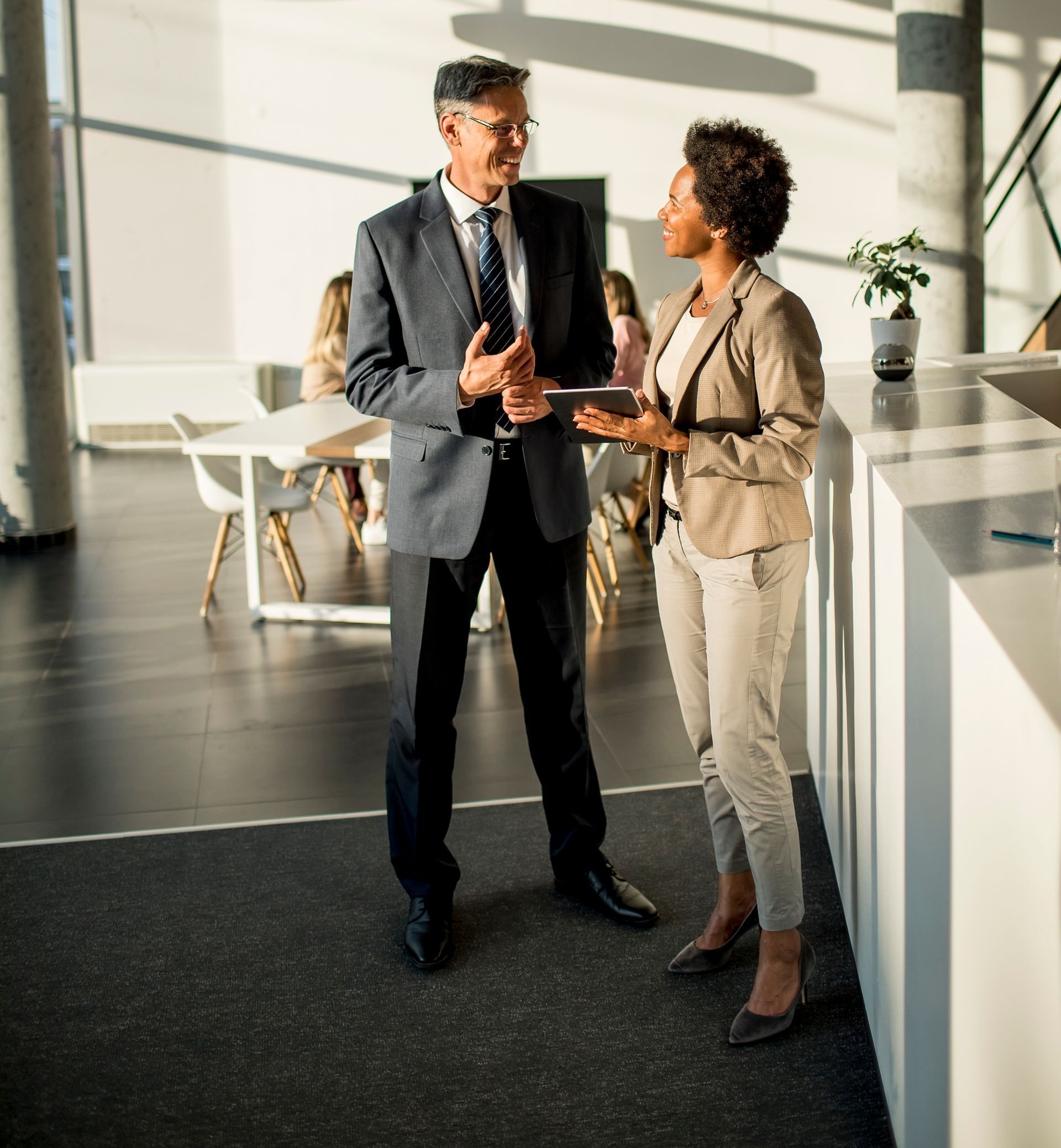 Two professional colleagues in business casual attire discuss work in a brightly lit, modern office lobby.