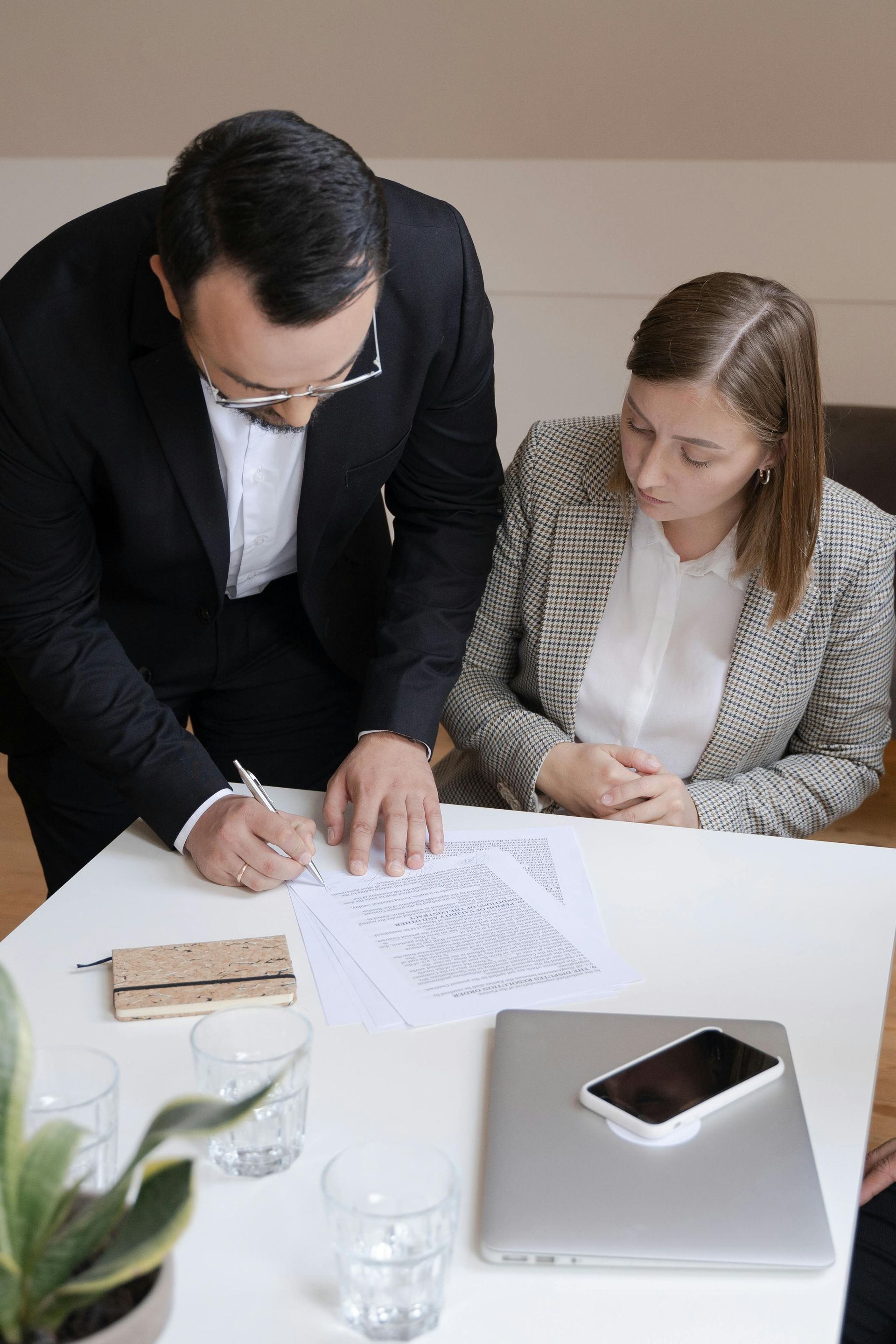 A person in a suit writes on a document as another looks on at a table with a laptop, phone, and glasses.