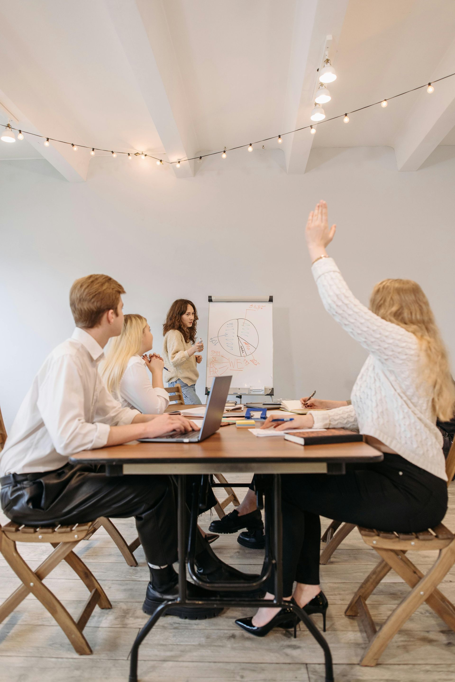 A group of people seated around a table in a bright office, with one person raising their hand during a presentation.