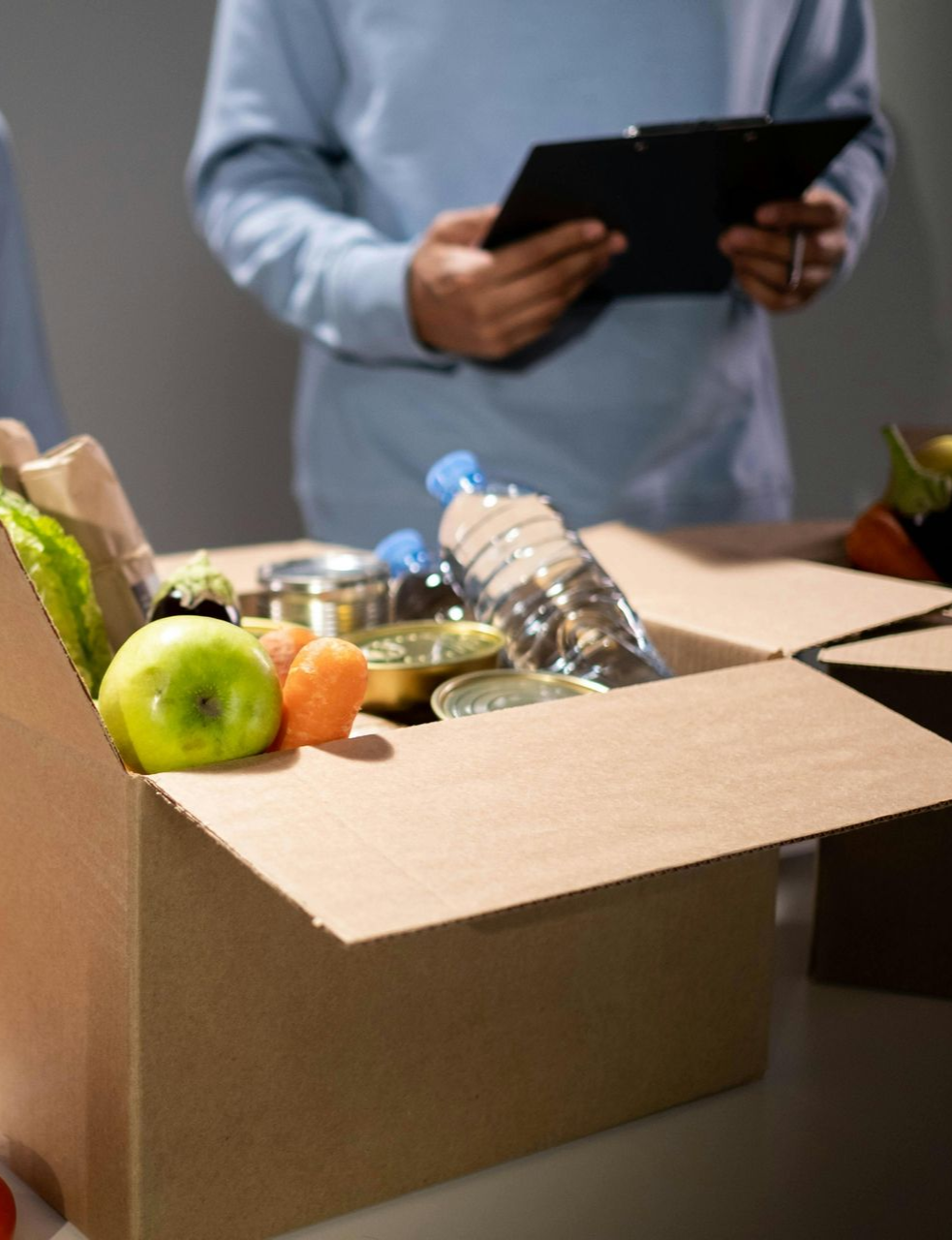 A person in a blue sweater holds a clipboard over a cardboard box filled with fresh produce and canned food.