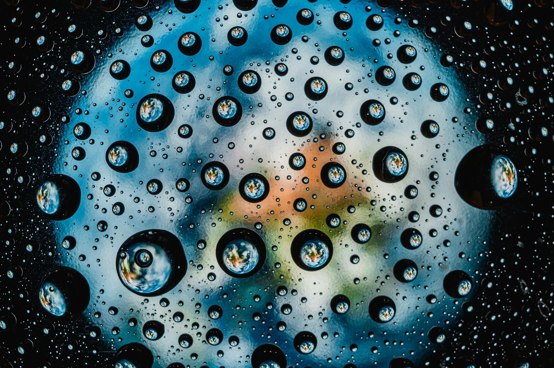 A close-up of water droplets on a surface, each acting as a tiny lens reflecting a colorful, blurred Earth-like image.