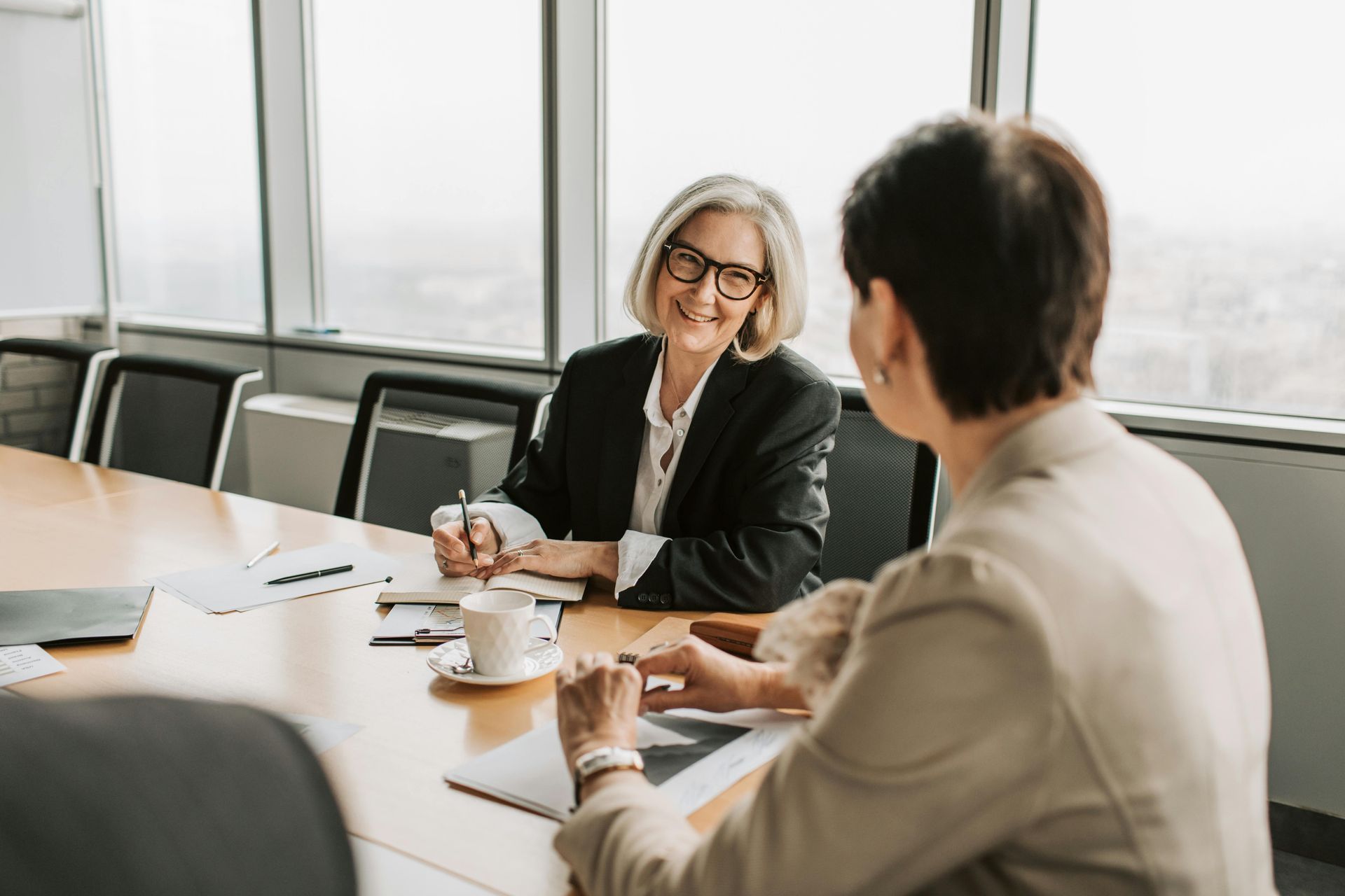 Two people in business attire meet at a conference table with documents and a coffee cup in a bright, modern office.