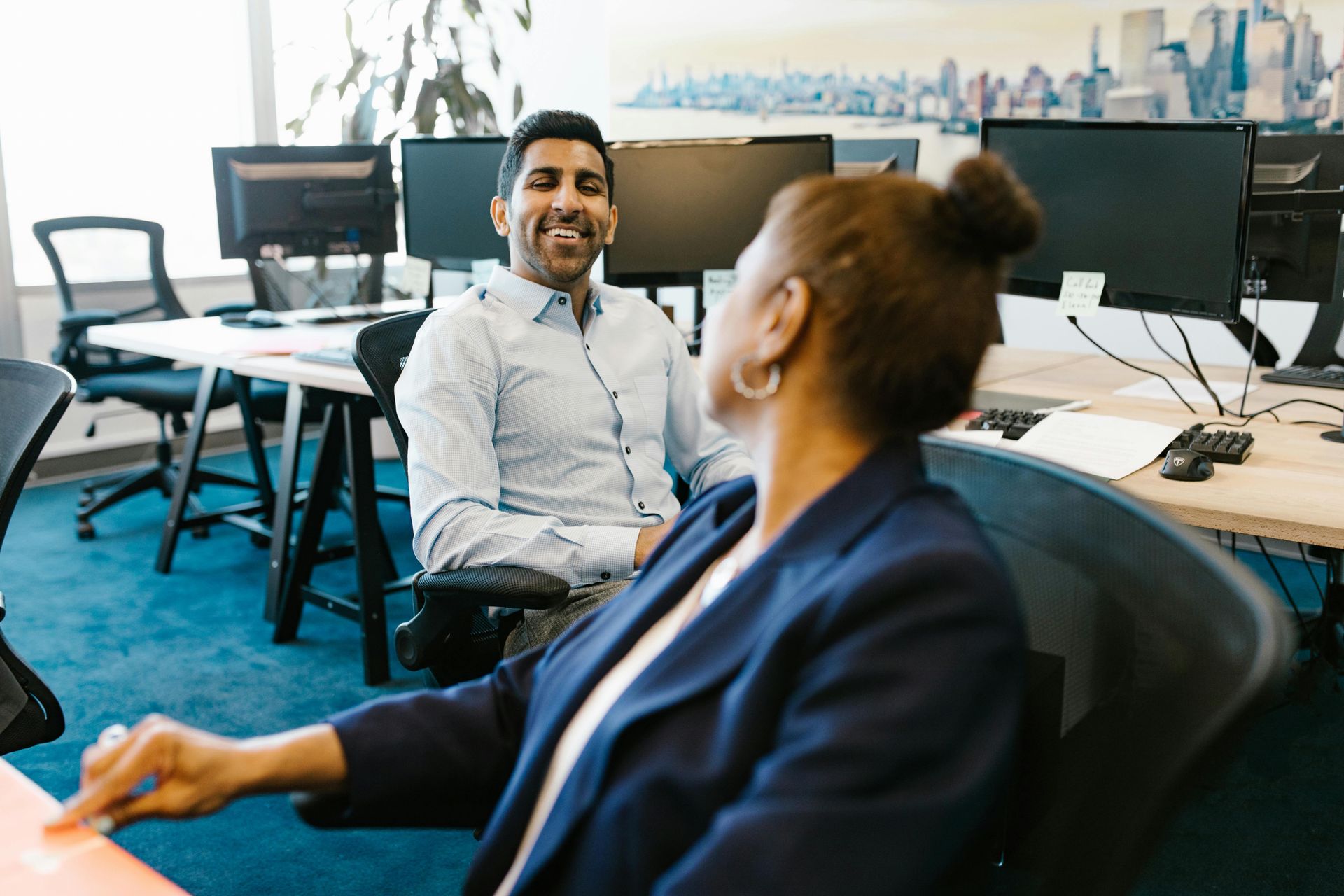 Two people in an office workspace are talking and looking at each other, with desks, computers, and chairs in the background.