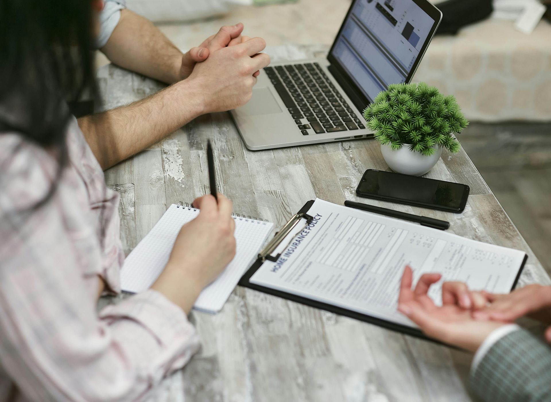 Two people discuss documents and a laptop at a table, one taking notes and the other gesturing.