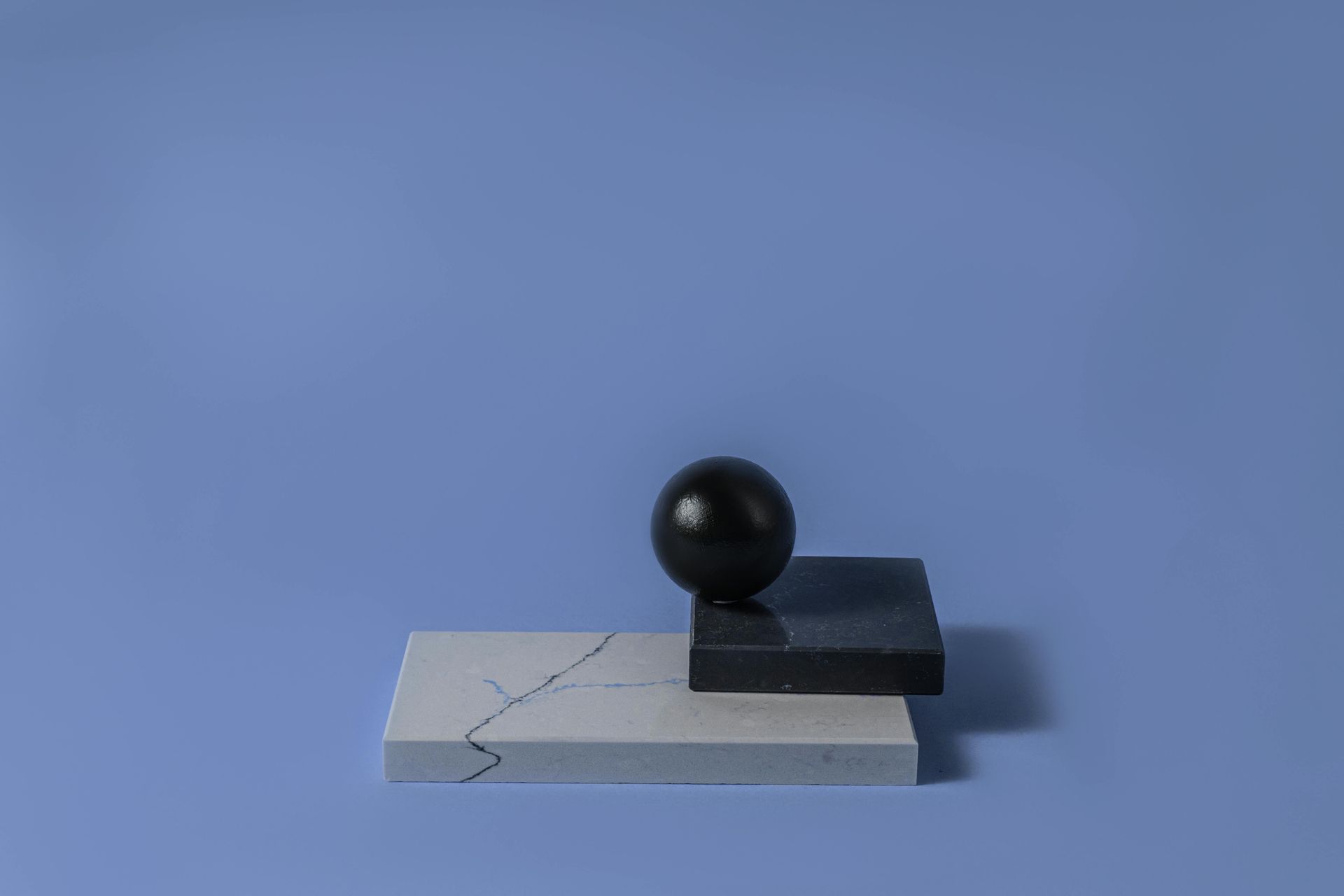 A black sphere rests on a small, dark rectangular block atop a larger, light gray rectangular base against a blue backdrop.