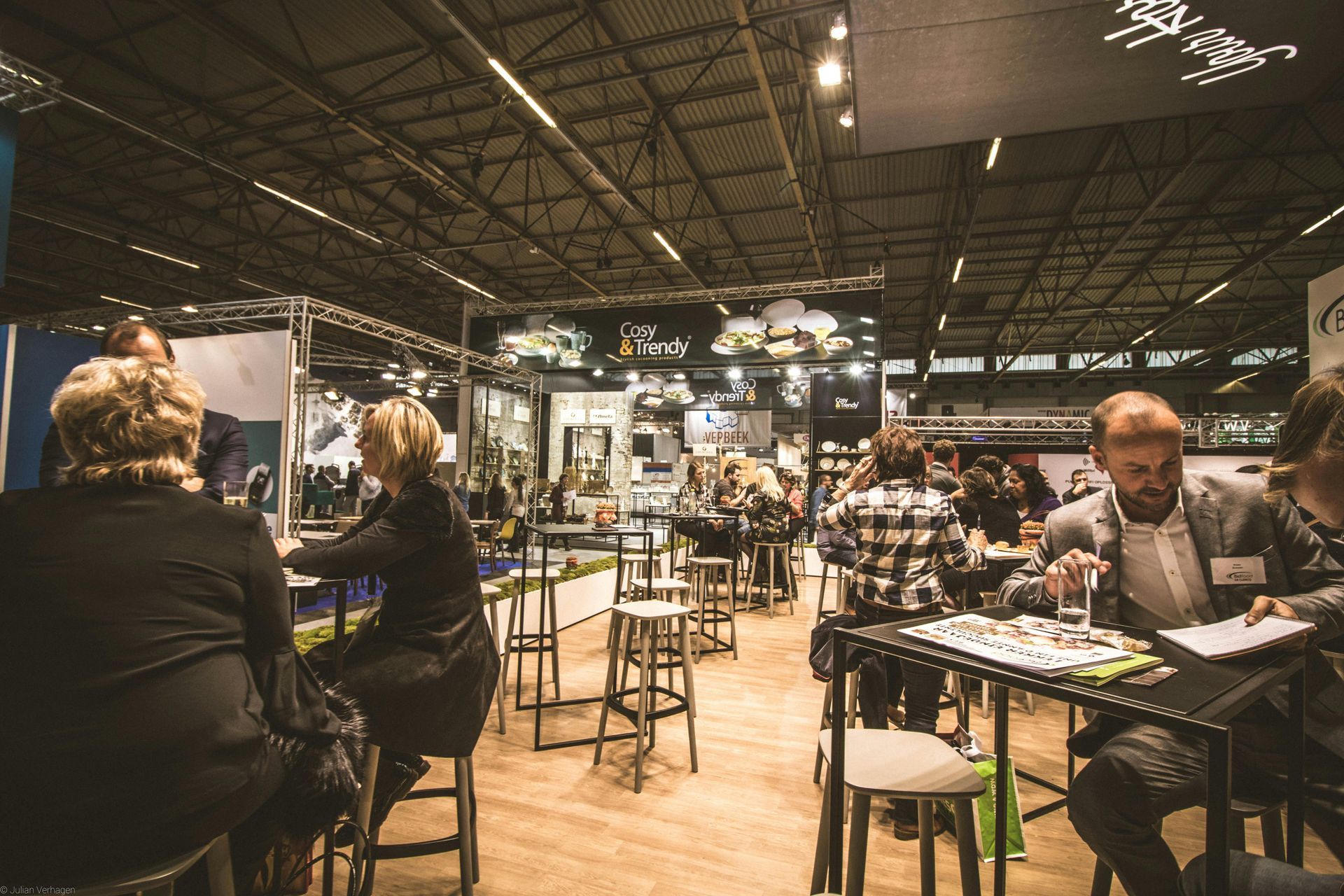 People seated at high tables in a bustling exhibition hall with overhead lighting and industrial-style rafters.