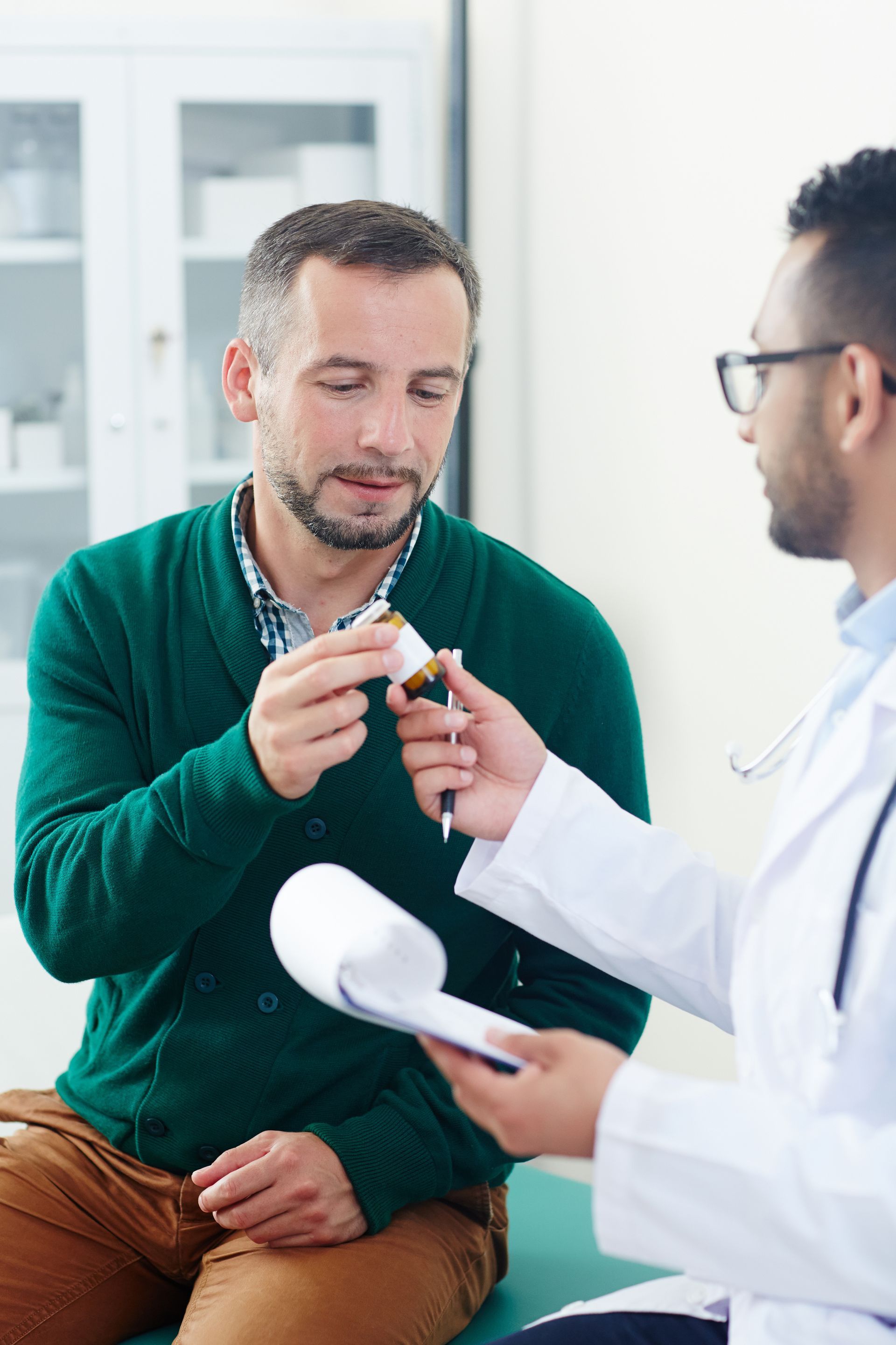 A doctor in a white coat shows a pill bottle to a patient during a medical consultation in an office.