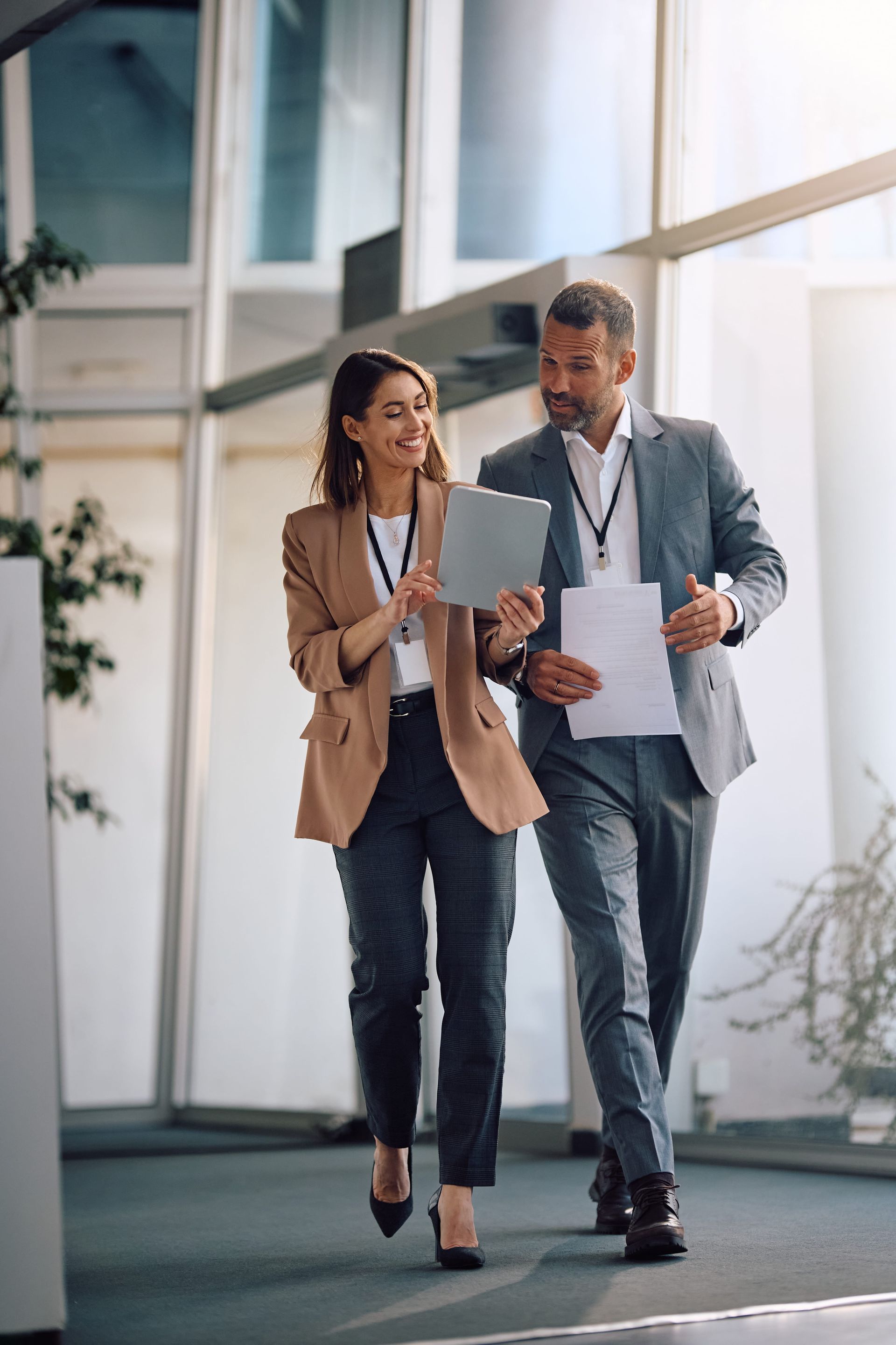 A professional person in a tan blazer and a person in a gray suit walk through an office, discussing a digital tablet.