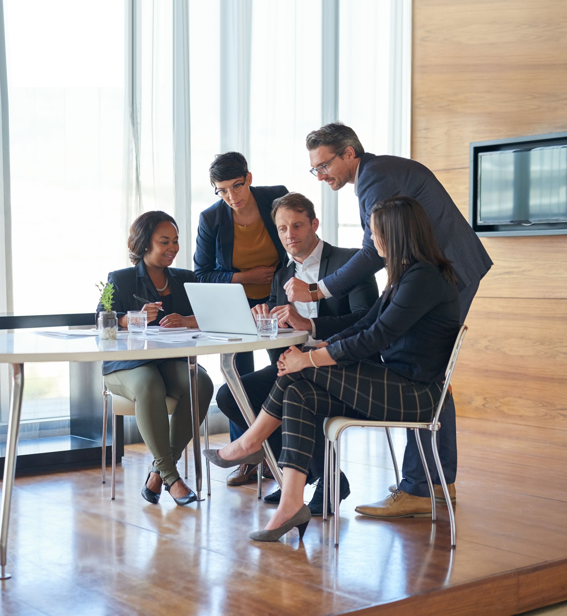 Four colleagues collaborate around a laptop on a white table in a bright, modern office with large windows.
